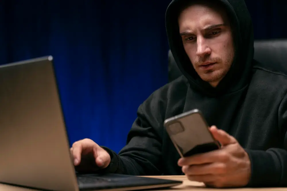 Homem de capuz preto em frente ao laptop sehurando o celular em uma mão com rosto preocupado.